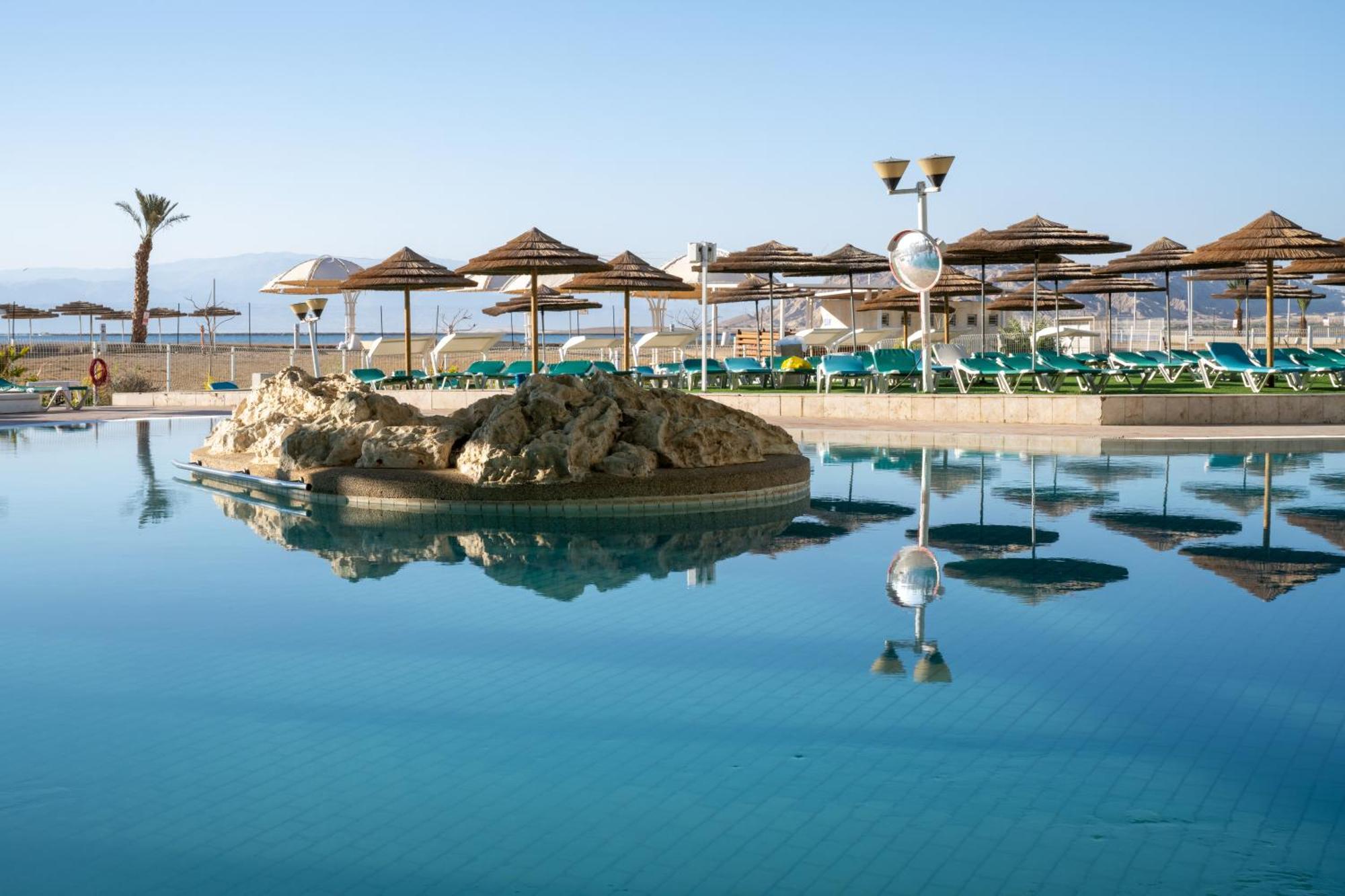 Leonardo Club Hotel Dead Sea Neve Zohar Exterior photo The photo shows a tranquil swimming pool area. It features a large, reflective pool with a small rock formation in the center. Surrounding the pool are several sun loungers with green cushions and a series of thatched umbrellas providing shade. In th