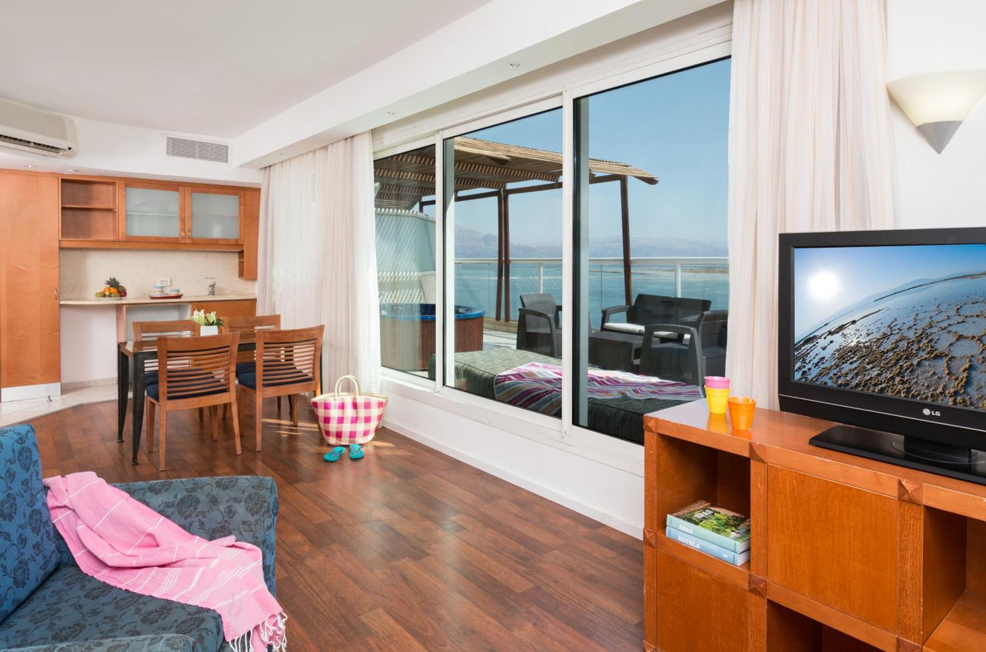 Leonardo Club Hotel Dead Sea Neve Zohar Exterior photo The photo depicts a modern living space with a view. On one side, there is a comfortable seating area with a couch and a television cabinet. The TV is turned on, showing a scenic image. 

In the background, there is a dining area with a wooden table 